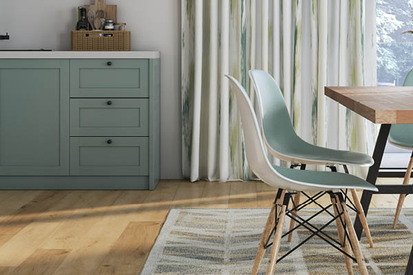 Ausschnitt eines Esszimmers mit hellgrüner Anrichte und passend hellgrünen Stühlen am Tisch. Auf dem Holzboden liegt unter dem Tisch ein Teppich und am Fenster hängt eine luftig weiße Gardine mit grünem Muster.